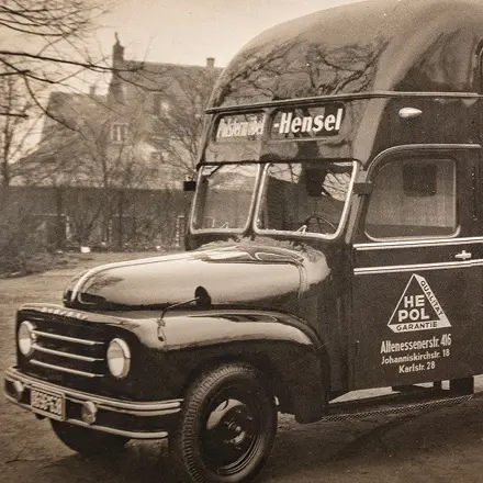 Schwarzweiss Foto eines Lieferwagens mit Aufschrift Möbel Hensel und HE POL Garantie, im Hintergrund Bäume und Häuser.